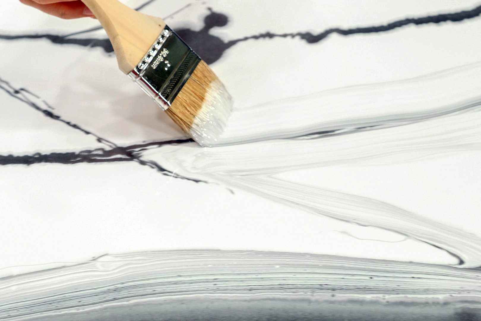 A hand is seen holding a paintbrush, applying light gray paint across a white canvas. Black lines intersect the canvas, creating an abstract design. The focus is on the brush and the painted strokes.
