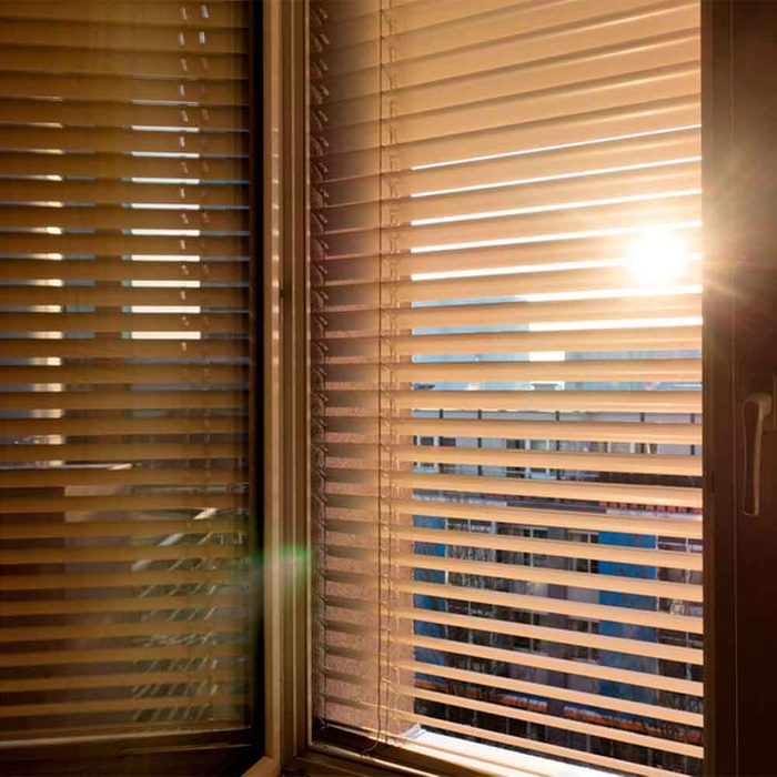 Blinds cover a window, partially allowing sunlight to stream through, illuminating a bright outdoor scene with buildings against a clear sky.