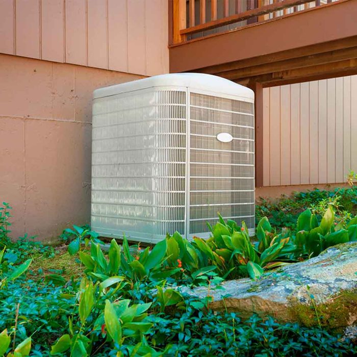 An air conditioning unit rests against a building, surrounded by green plants and rocks, providing cooling in a residential outdoor environment.