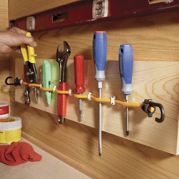 A hand organizes various tools hung on a wooden panel, using a yellow rope and clips for storage, in a workshop setting.