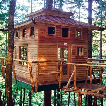 A wooden treehouse stands elevated among trees, featuring multiple windows and a wraparound balcony, creating a cozy retreat in a natural forest setting.