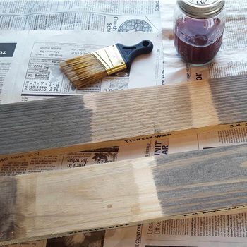 Wooden boards display various stain applications, partially finished. A paintbrush rests on newspaper beside a jar of red stain, indicating a woodworking project.