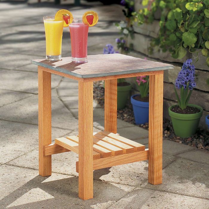 Two colorful drinks sit on a wooden table with a slate top in a sunny outdoor setting, surrounded by potted plants and a stone wall.
