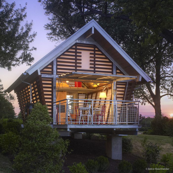 A modern house with a sloped roof showcases a welcoming interior, illuminated by warm lights, surrounded by trees and shrubs at sunset.