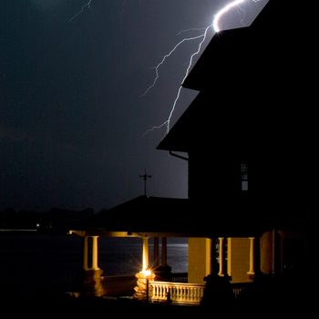A house stands in darkness, illuminated by a nearby porch light, as lightning strikes in the night sky, highlighting the stormy atmosphere.