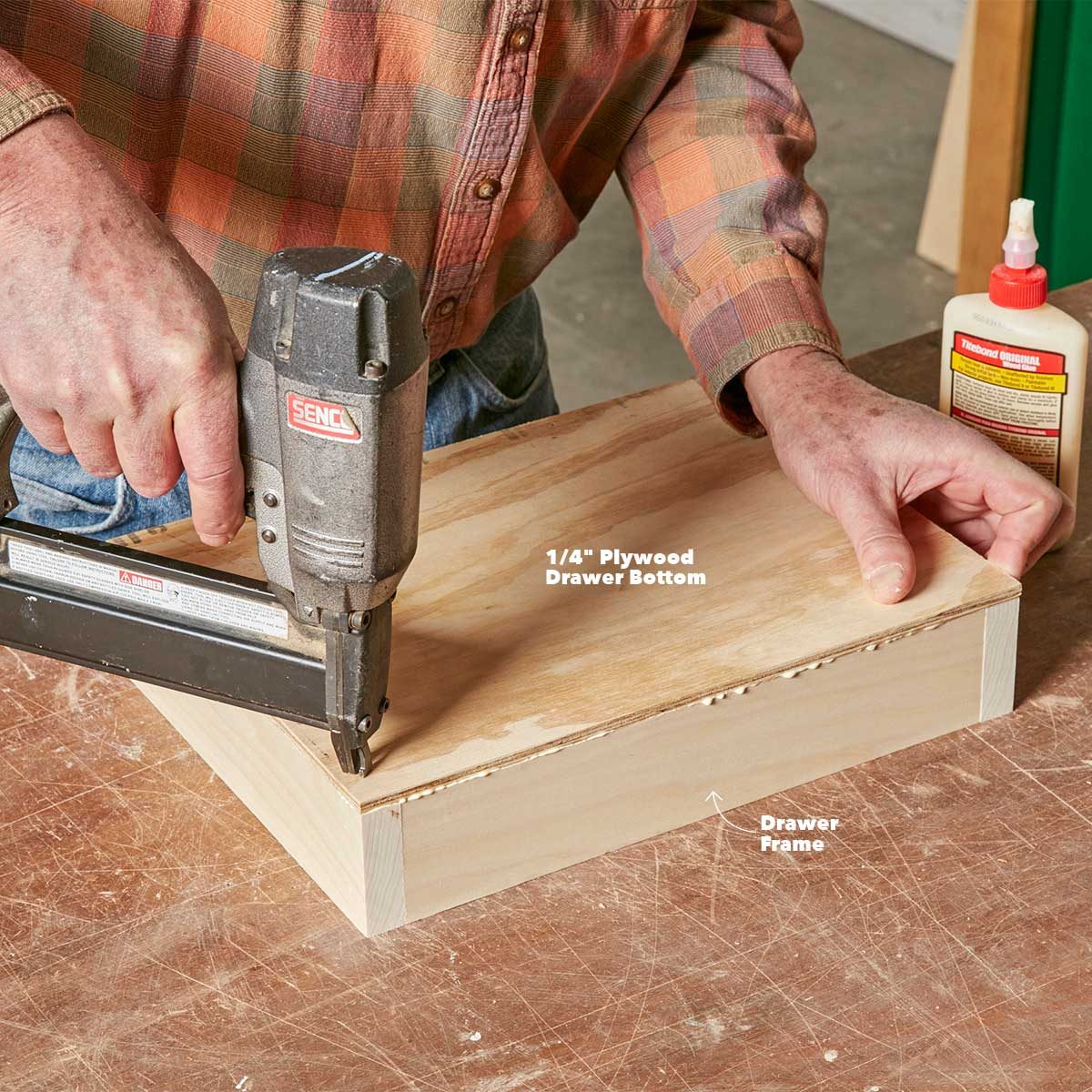 A person uses a nail gun to attach a plywood panel to a wooden drawer frame on a work surface, with wood glue visible nearby.