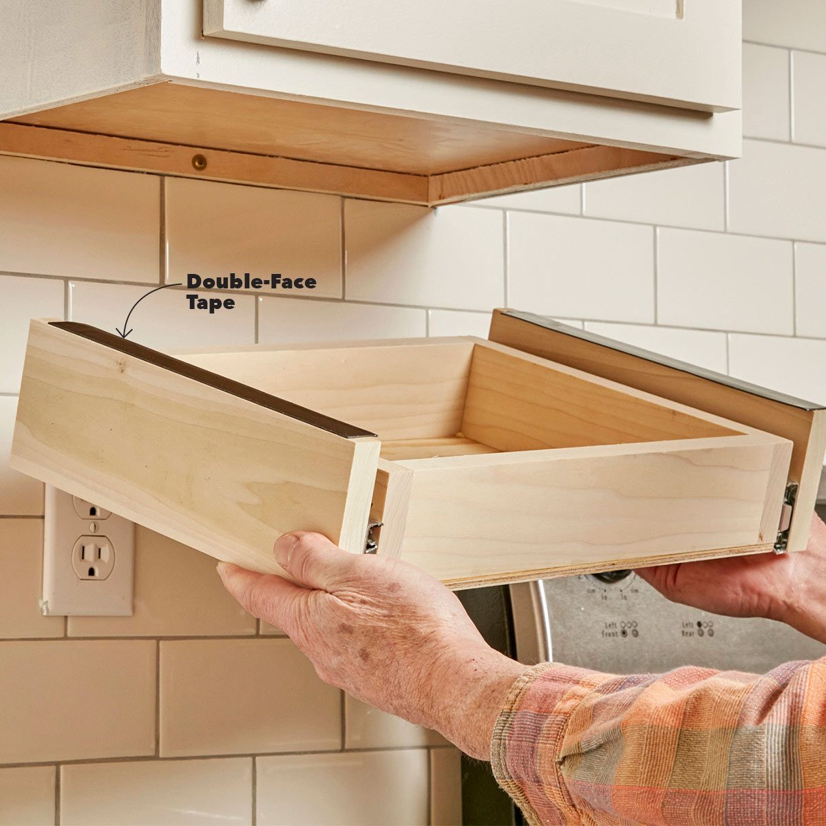 A wooden drawer is being held up for installation, with double-face tape indicated, against a tiled kitchen backdrop and an electrical outlet visible below.