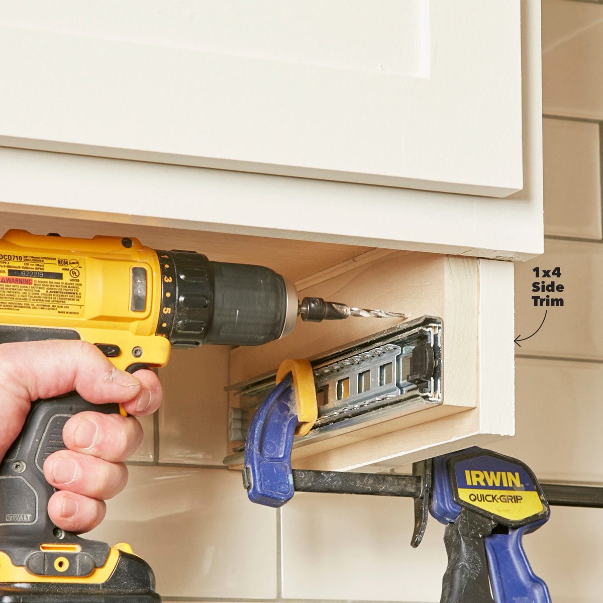 A hand holds a drill, attaching a side trim to a cabinet while clamps secure the trim in place against a tiled backdrop.