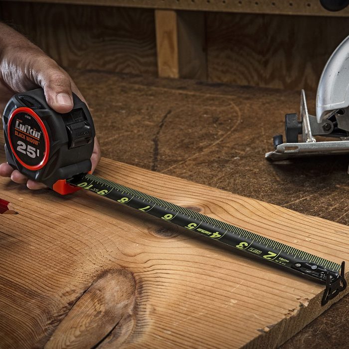 A tape measure extended over a wooden plank, with a hand holding it in place on a workbench, alongside a saw and red pencil.