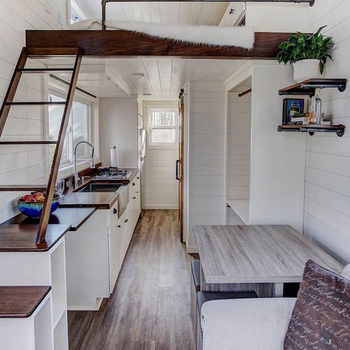 A wooden ladder leads to a lofted bed above a compact kitchen with white cabinets. A dining table and cozy seating occupy the living area.