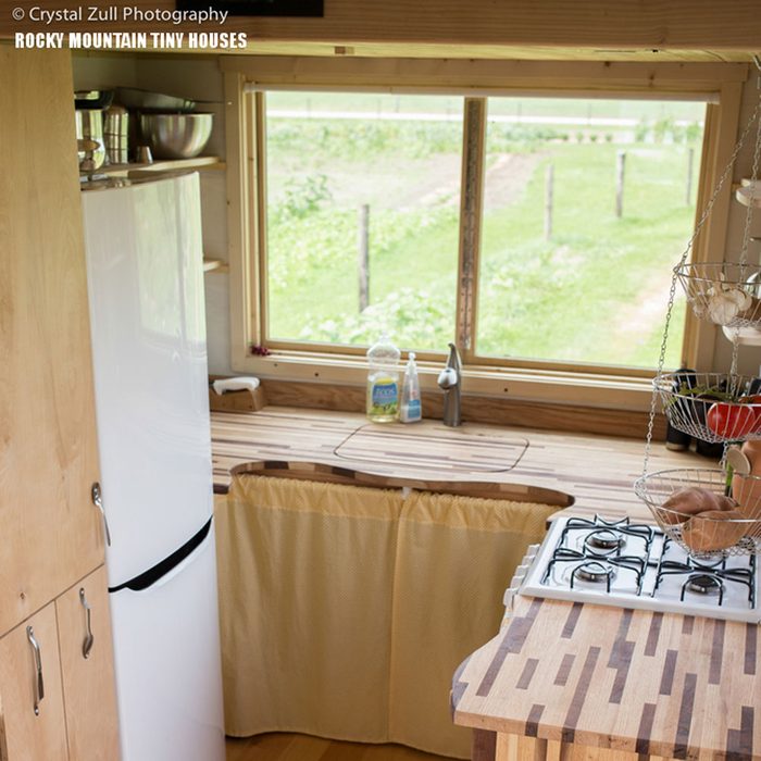 A kitchen features a white fridge, wooden countertops, and a sink. Sunlight streams through a window, revealing a green garden outside.