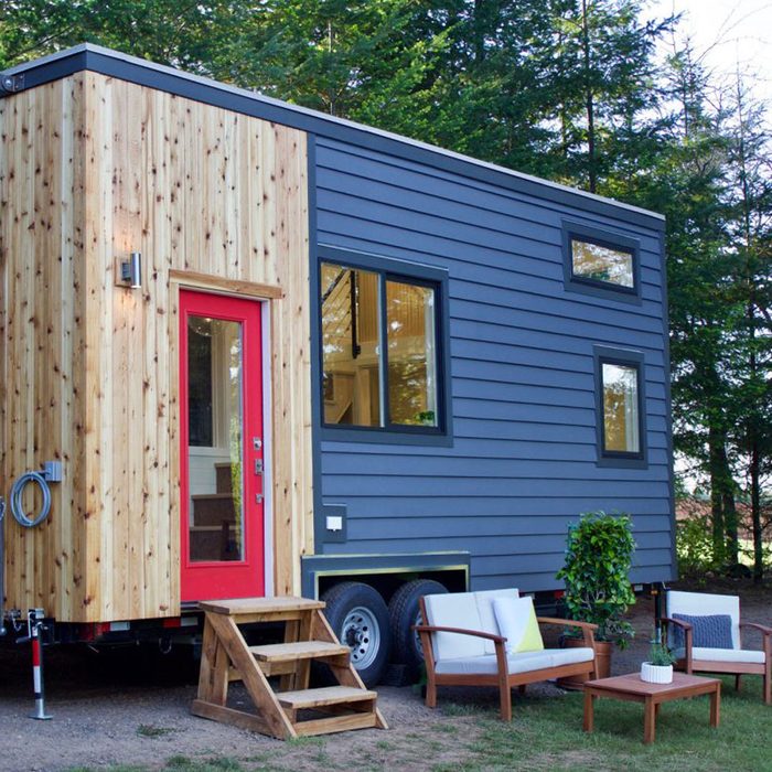 A tiny house with a wooden and gray exterior features a bright red door, steps, and outdoor seating surrounded by trees in a natural setting.