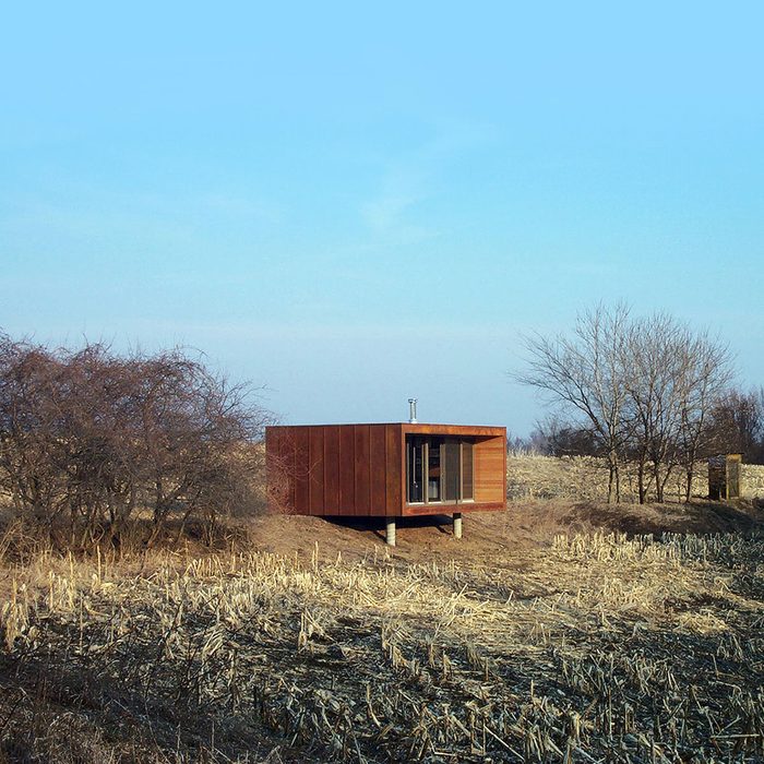 A modern wooden cabin stands elevated in a barren field, surrounded by sparse trees under a clear blue sky.
