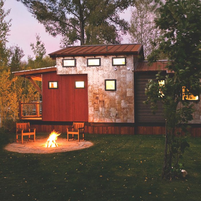 A rustic cabin stands beside a circular fire pit, with flames flickering. Two wooden chairs face the fire, surrounded by grassy landscaping and trees.