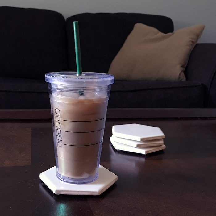 A clear cup with ice coffee sits on a white hexagonal coaster, beside stacked hexagonal coasters, in a living room with a dark couch.