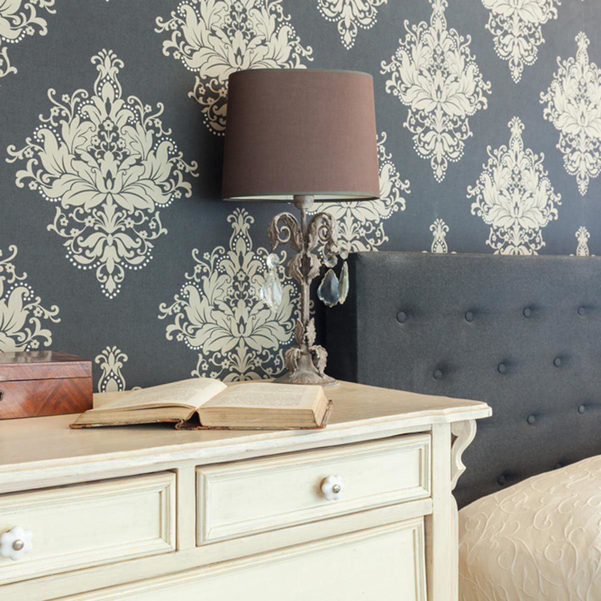 A decorative lamp stands on a dresser beside an open book, against a richly patterned wallpaper, featuring a tufted bedhead in the background.
