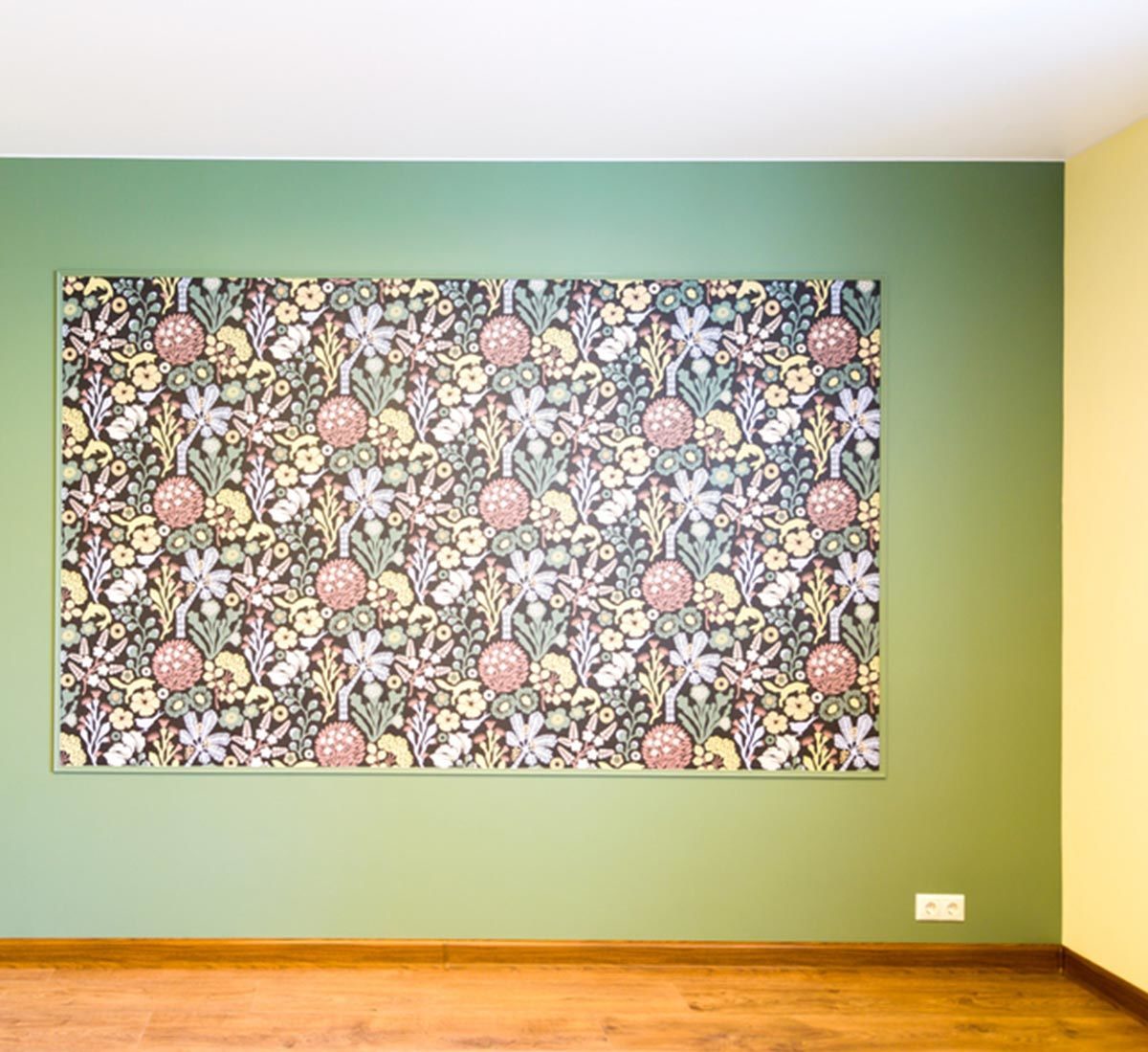 A large floral wallpaper is hung on a green wall within a room, contrasting with a yellow wall adjacent, and wooden flooring below.