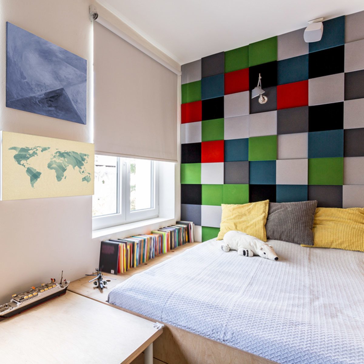 A bed with colorful pillows sits against a patterned wall. Nearby, a wooden desk displays a ship model, while a window lets in natural light.