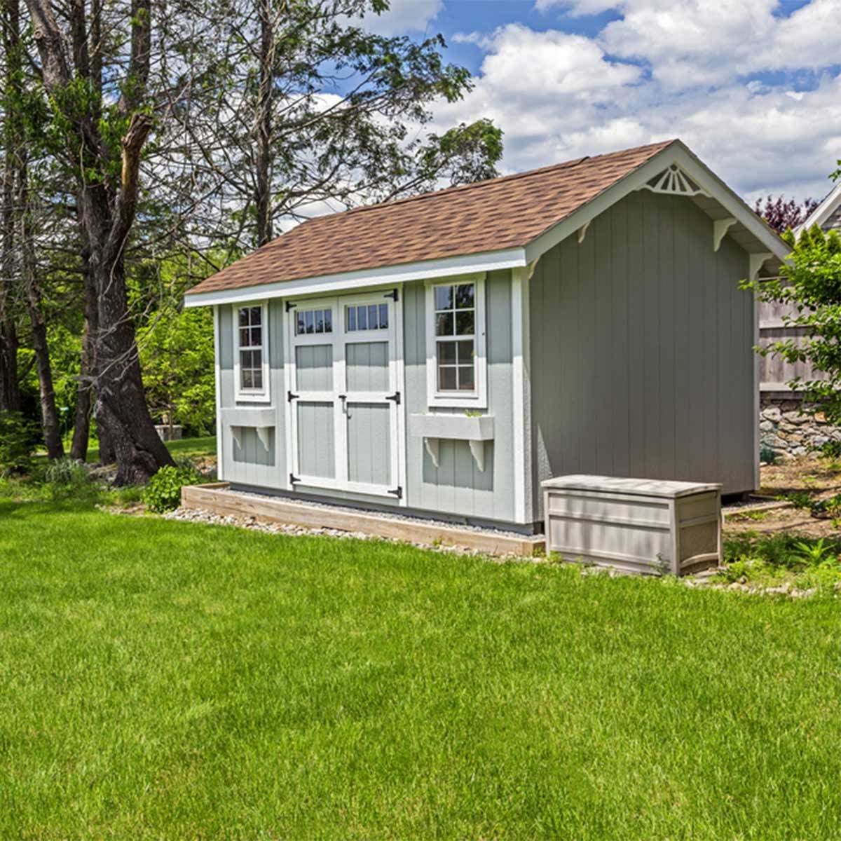 A small, light-colored shed sits on a grassy lawn, with trees in the background and a storage box nearby under a clear blue sky.