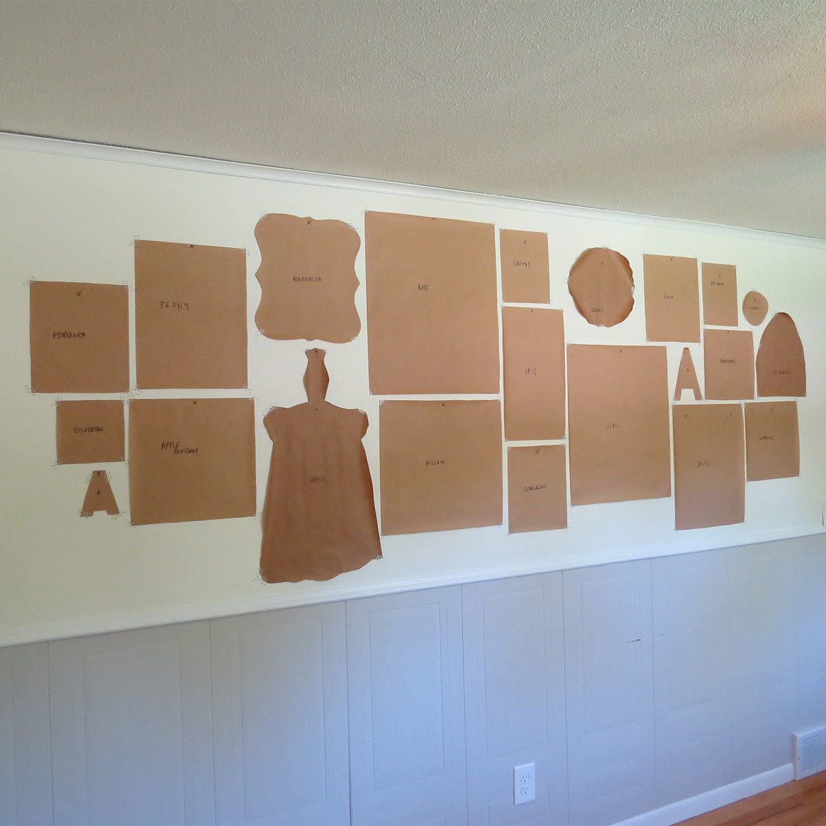 Brown paper patterns are pinned to a wall, each labeled with letters or words, in a well-lit, minimalist room with light-colored walls and wooden flooring.