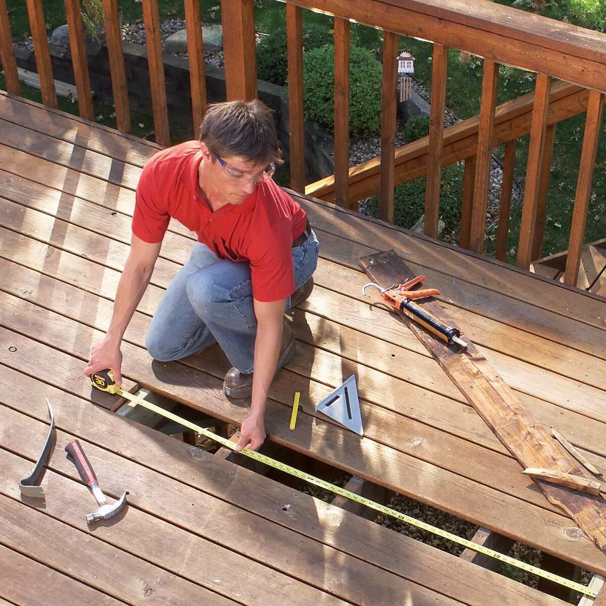 A man kneels on a wooden deck, measuring a gap with a tape measure, surrounded by tools and wooden planks in a backyard setting.