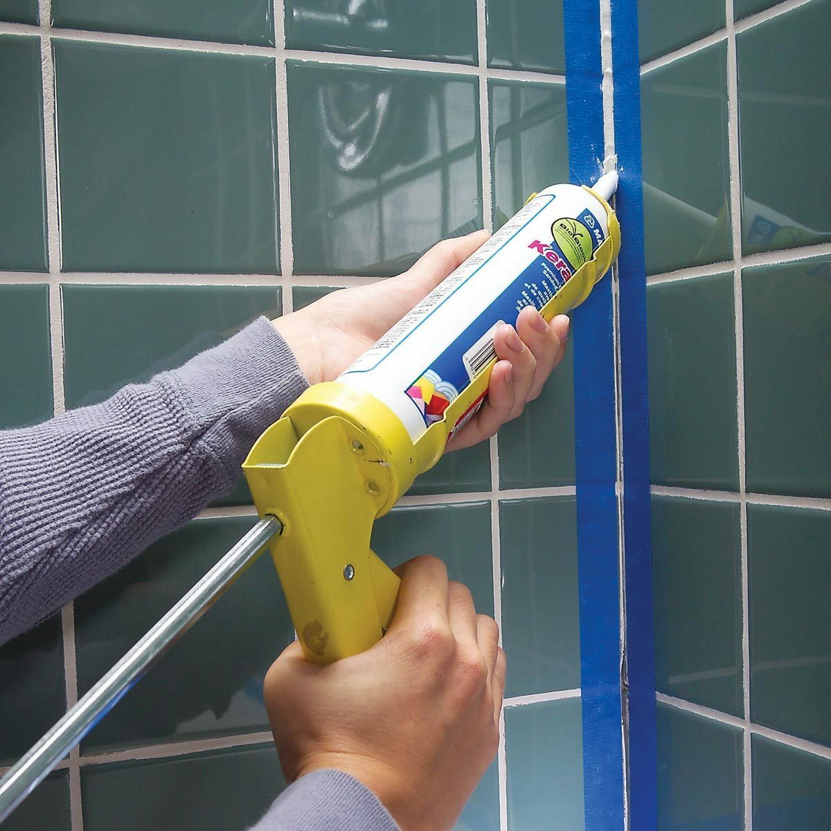 A person uses a caulking gun to apply sealant along a blue tape line on tiled green wall, in a bathroom setting.