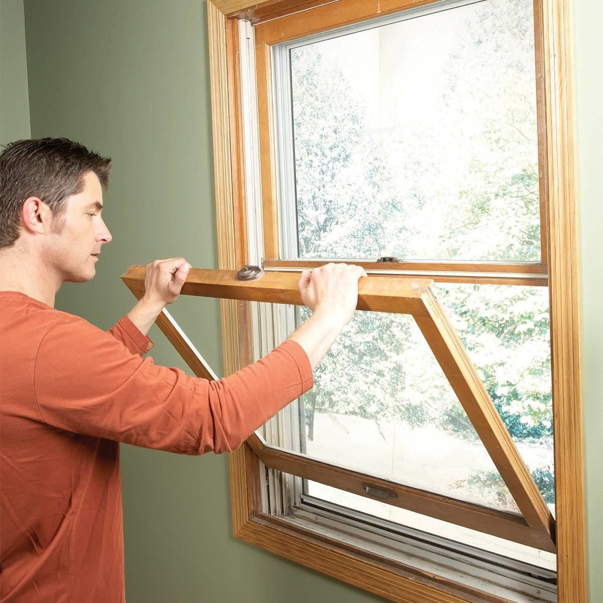 A man tilts a wooden window open, revealing a green outdoor landscape through a partially transparent pane, set against a light green wall.