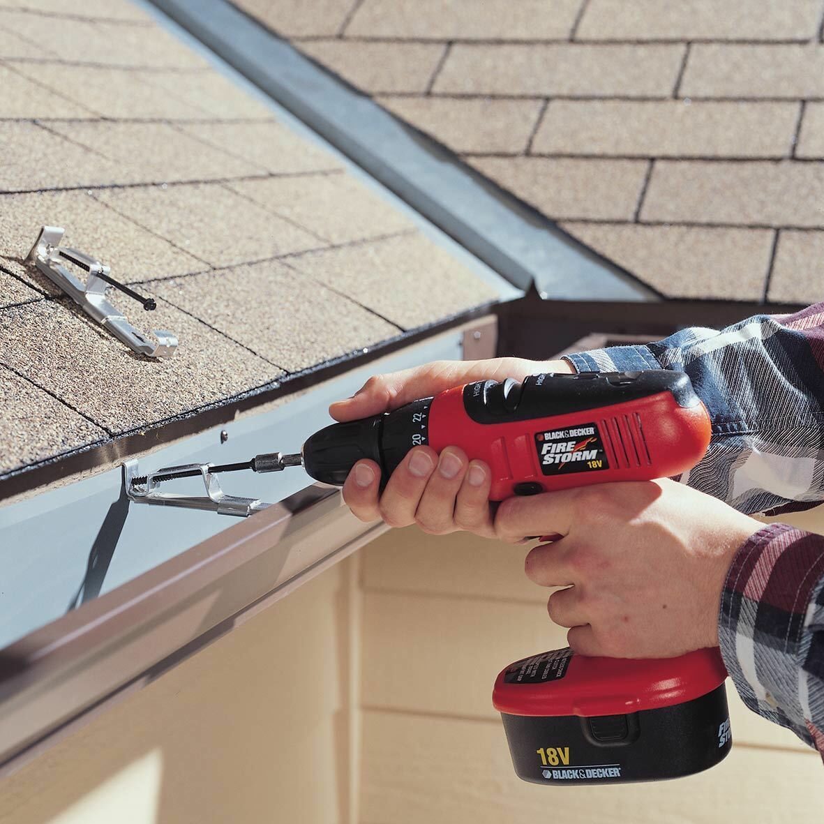 A person is using a cordless drill to secure a bracket onto a metal gutter, with shingled roofing in the background and sunlight illuminating the scene.