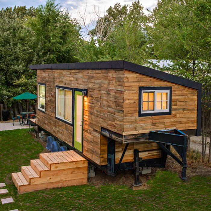 A wooden tiny house stands elevated on supports, with steps leading to the door, surrounded by grass and trees in a tranquil outdoor setting.