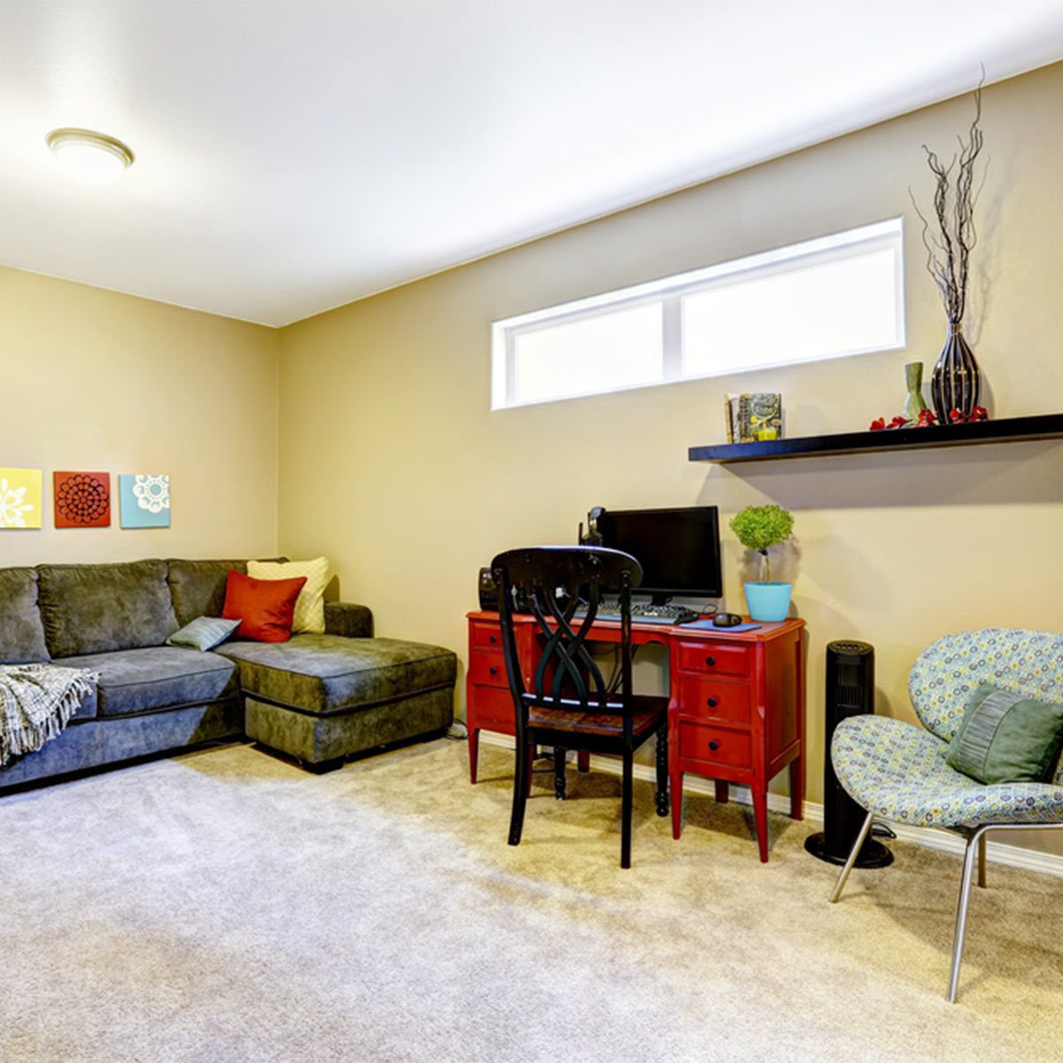 A gray sectional sofa faces a red desk with a computer in a cozy room, adorned with wall art and a decorative shelf.