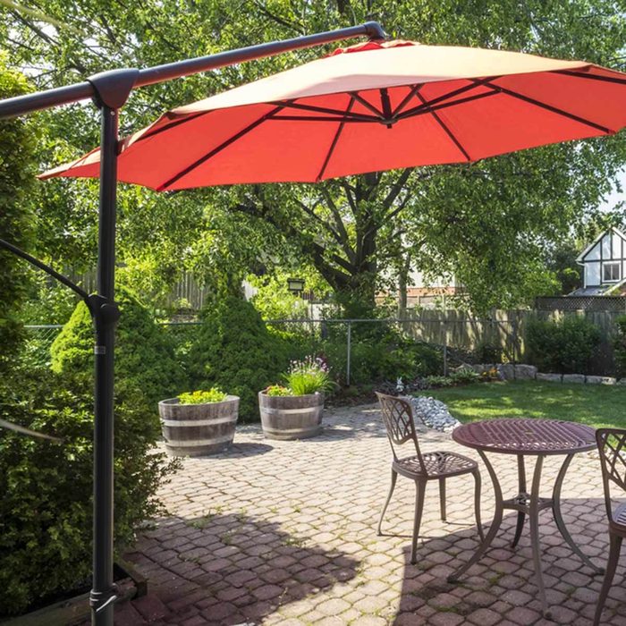 A red patio umbrella stands upright, shading a small round table and chair on a stone-paved yard surrounded by lush greenery and flower pots.
