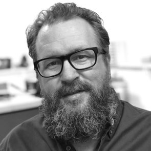 A man with glasses smiles, showcasing a thick beard, against a blurred background suggesting an office setting.