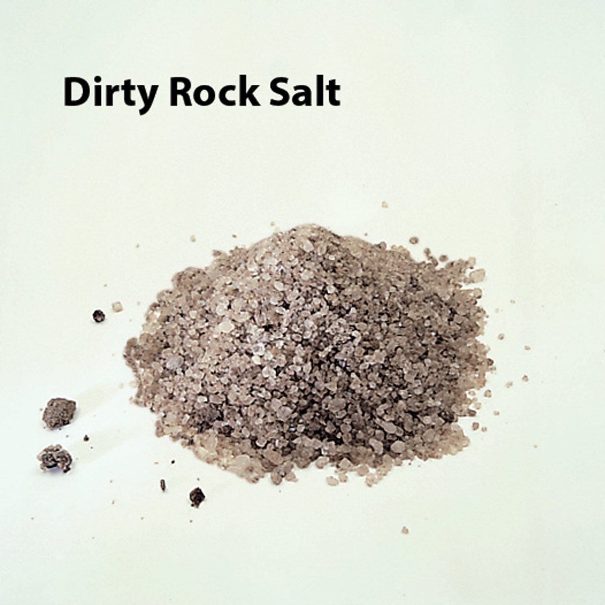 A mound of dirty rock salt rests on a light background, with small, irregular grains and darker specks visible in the pile.