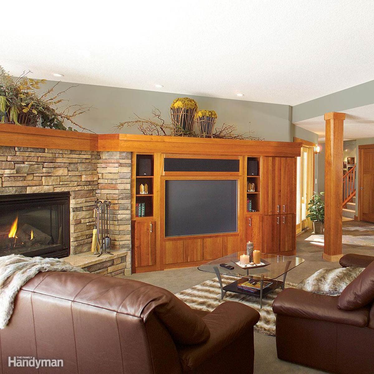 A cozy living room features leather couches facing a stone fireplace, with wooden cabinetry and a glass coffee table, illuminated by overhead lighting.