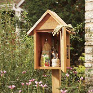 A garden shed stands on a post, displaying gardening tools inside. Pink flowers grow nearby, with greenery providing a natural backdrop. A pair of gloves rests on top.