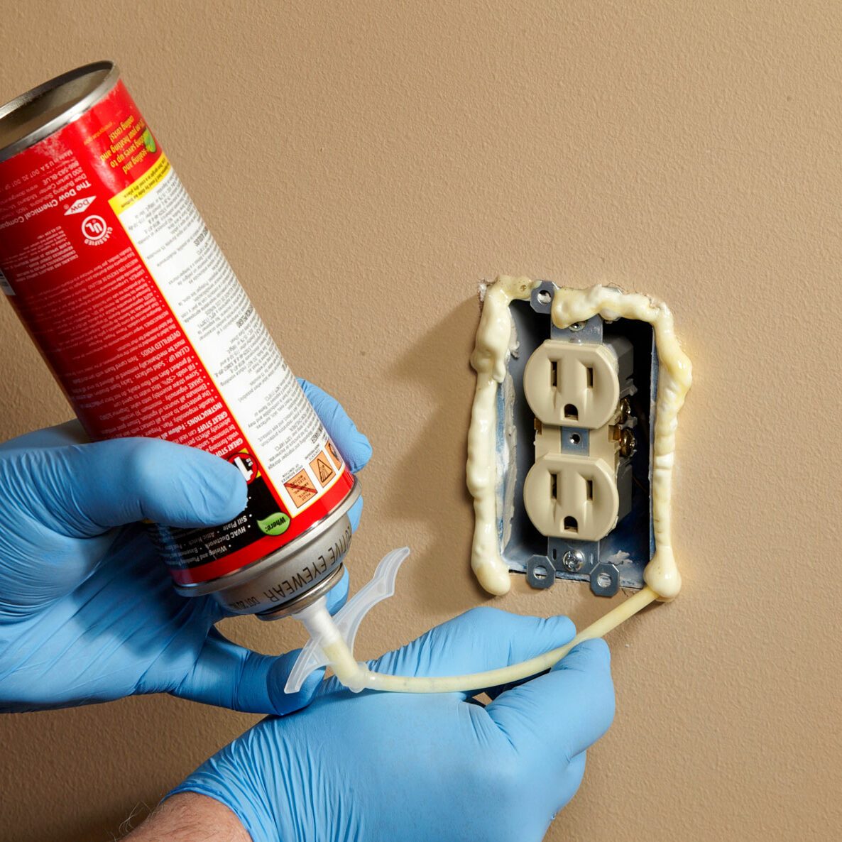 A hand in a blue glove sprays foam insulation into an electrical outlet on a beige wall, sealing gaps around the outlet.