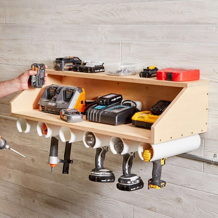 A wooden shelf holds various power tools and batteries, while a hand reaches for a tool, positioned against a textured wall.