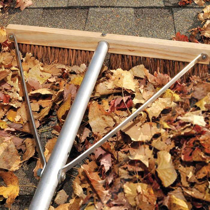 A large, metal rake is sweeping fallen, colorful autumn leaves off a rooftop, with a textured, dark surface beneath the leaves.
