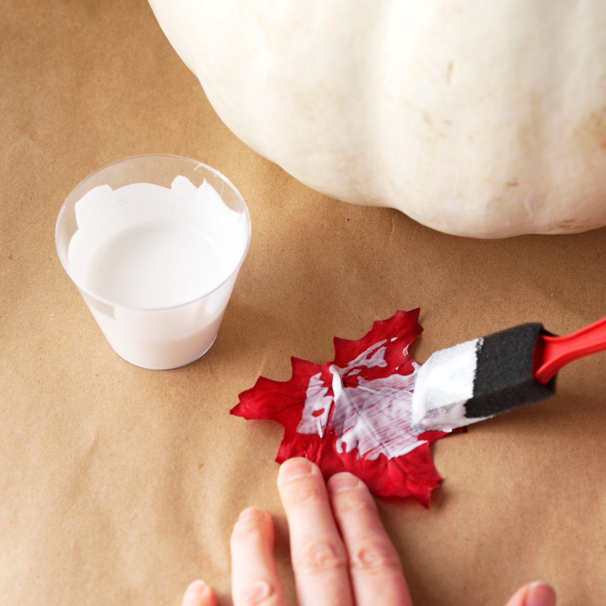 A hand applies white glue to a red leaf using a foam brush, surrounded by a clear cup of glue and a large white pumpkin on brown paper.