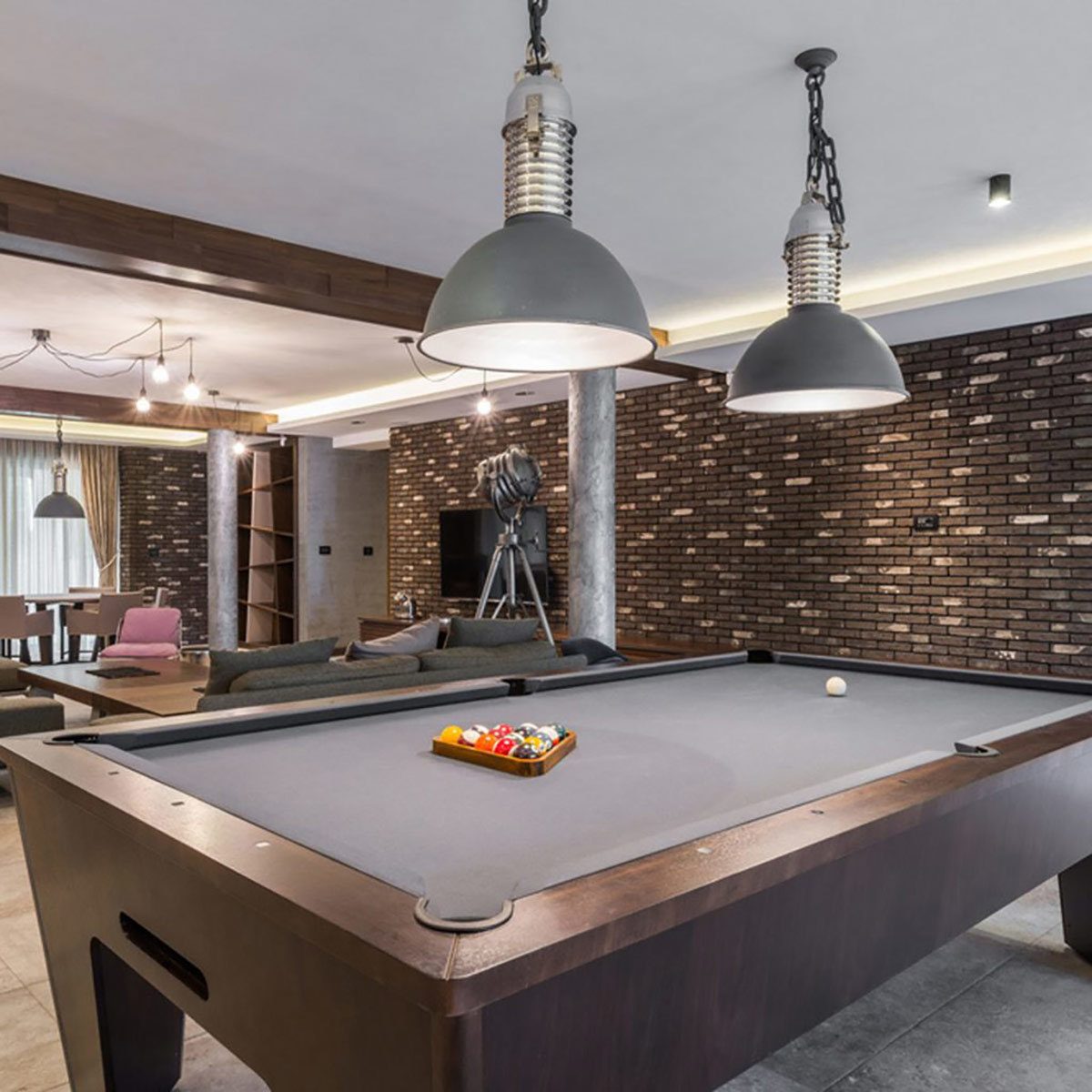 A billiard table holds colored balls, while pendant lights illuminate a modern lounge. A brick wall and sleek furnishings complete the stylish environment.