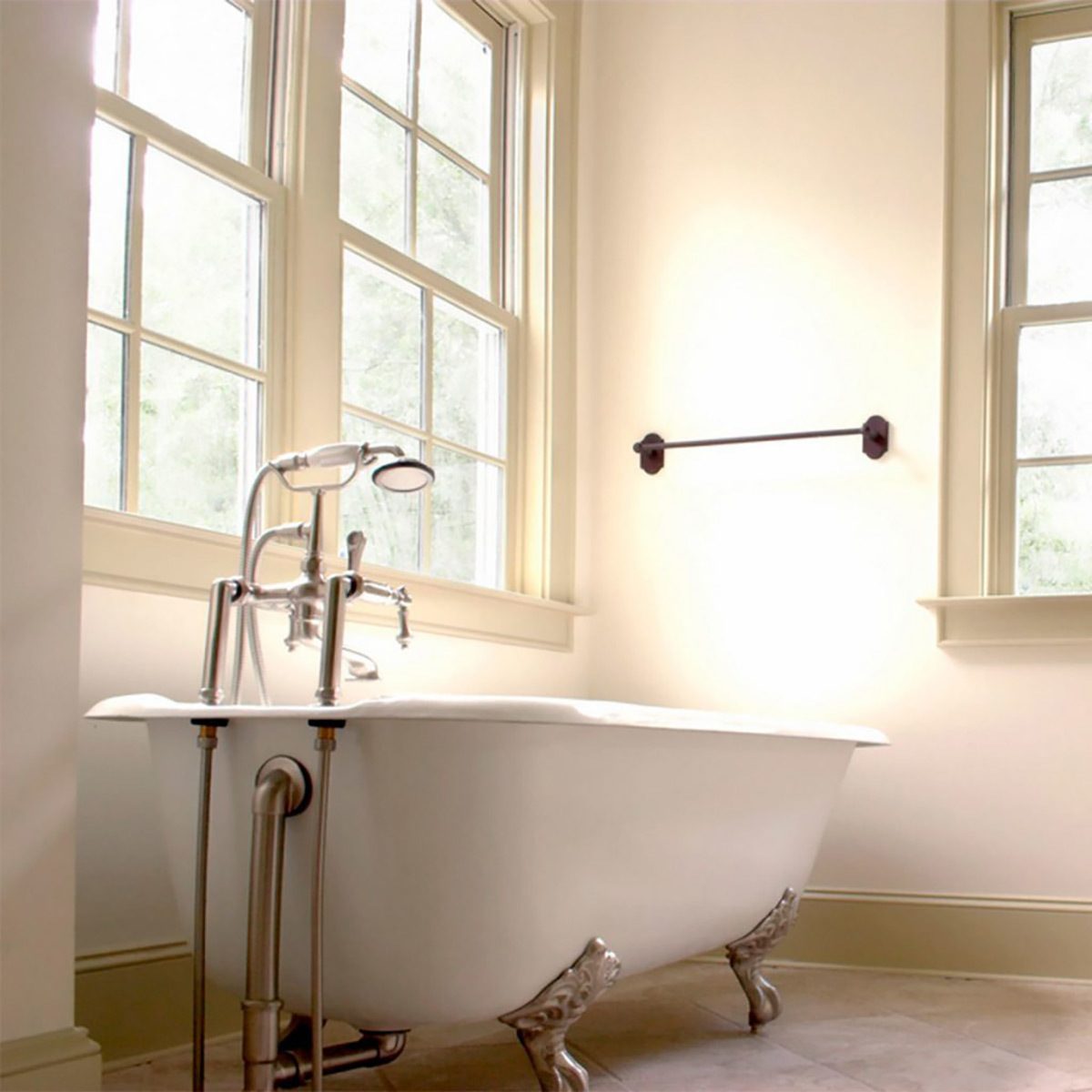 A freestanding white bathtub with ornate metal legs sits in a bright bathroom, beside large windows that provide natural light.