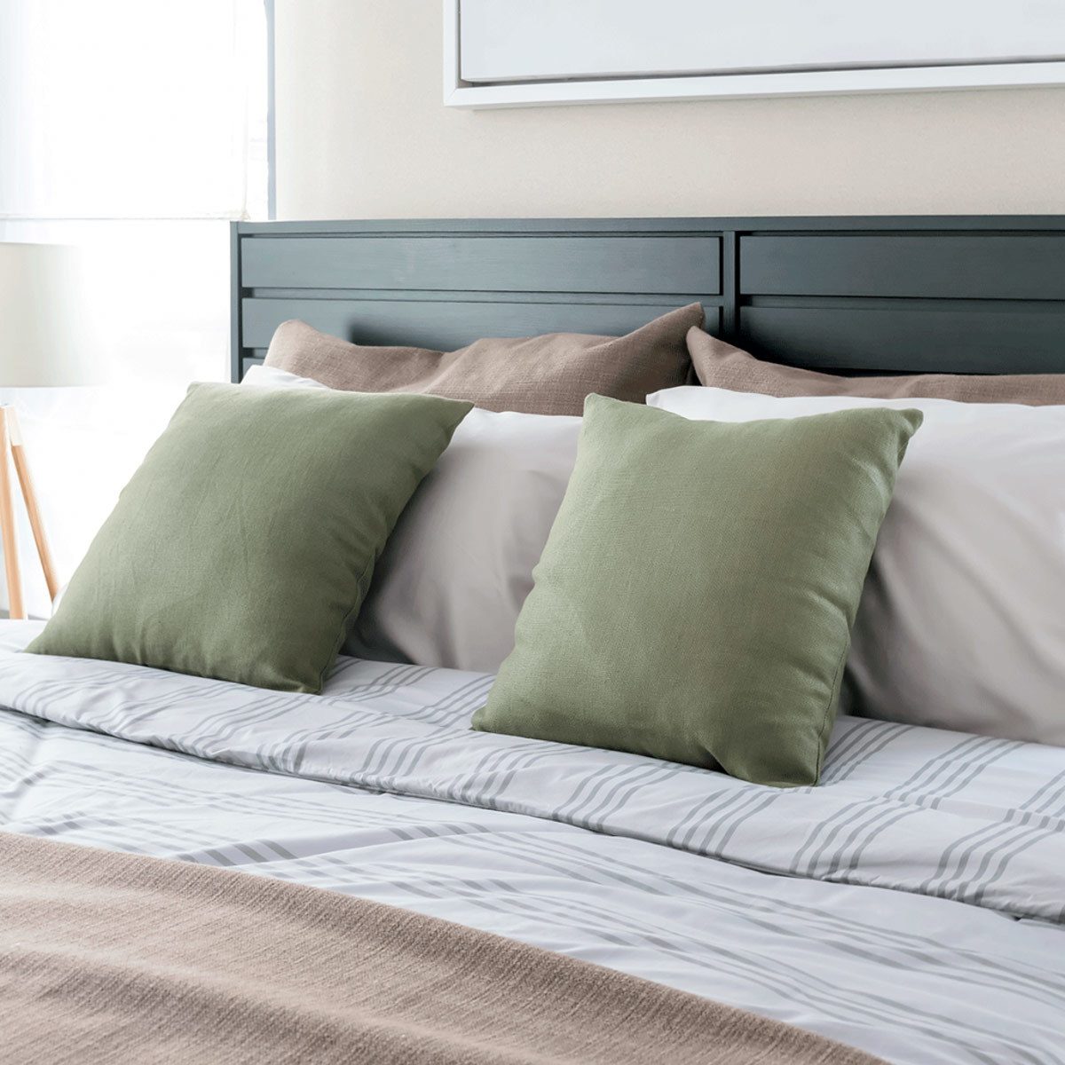 Two green pillows are positioned on a neatly made bed, accompanied by a light brown throw and striped bedding, in a bright, softly lit room.