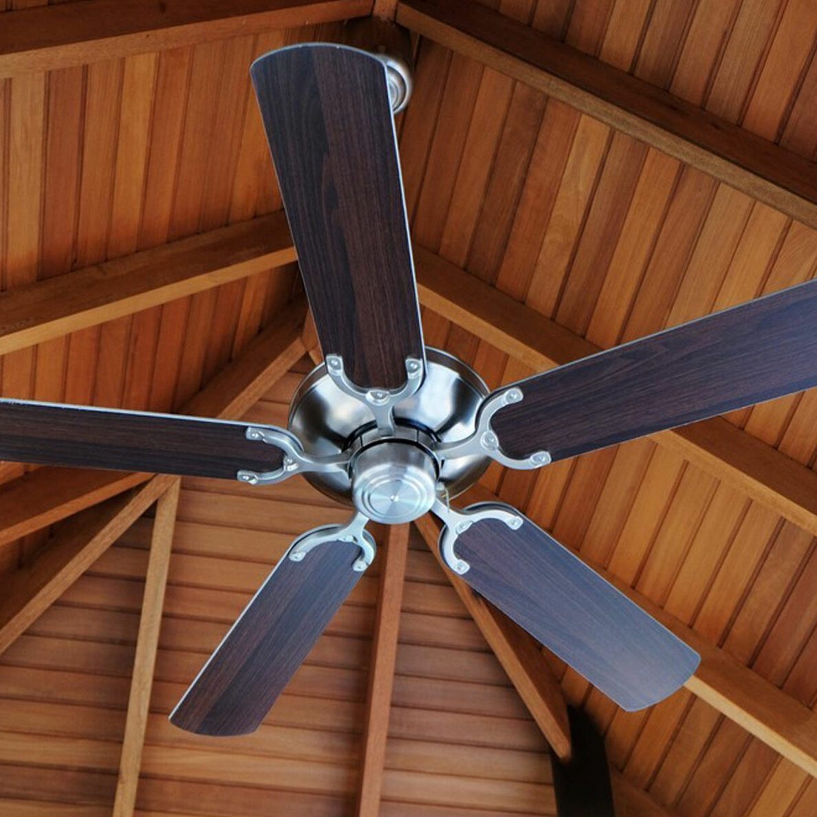 A ceiling fan with five wooden blades spins, suspended from a wooden beam ceiling, creating airflow in a cozy interior space.