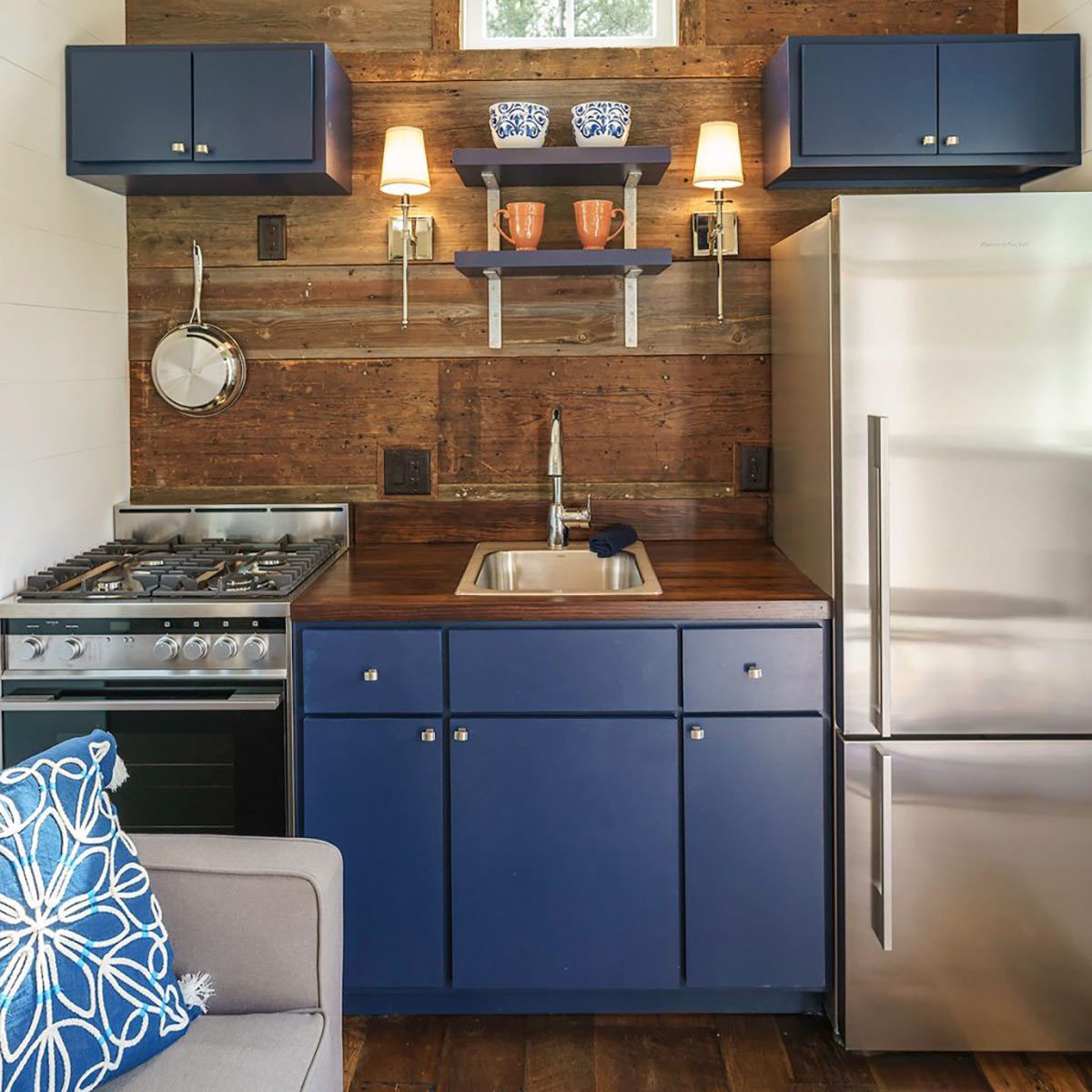 A modern kitchen features a stove, sink, and refrigerator, with wooden walls, blue cabinets, and decorative dishware on shelves, creating a cozy atmosphere.