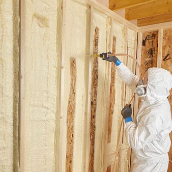 A person in protective gear sprays insulation foam onto a wooden wall framework, working indoors within an unfinished building.