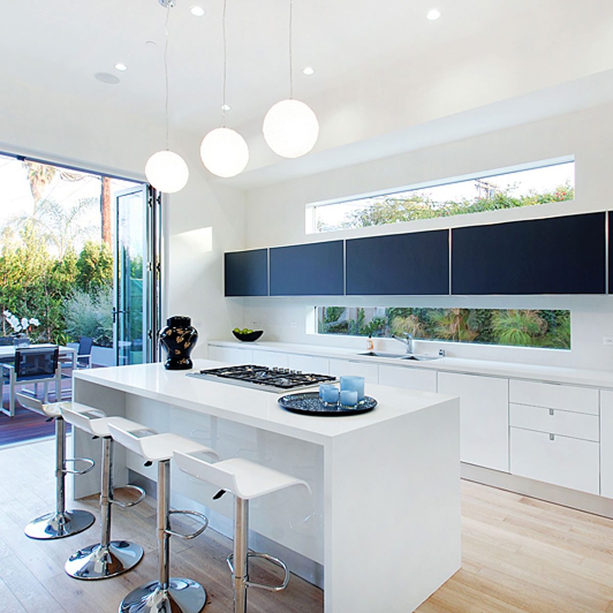 A modern kitchen features a white island with bar stools, a black stove, and large windows revealing a lush outdoor garden. Three pendant lights illuminate the space.