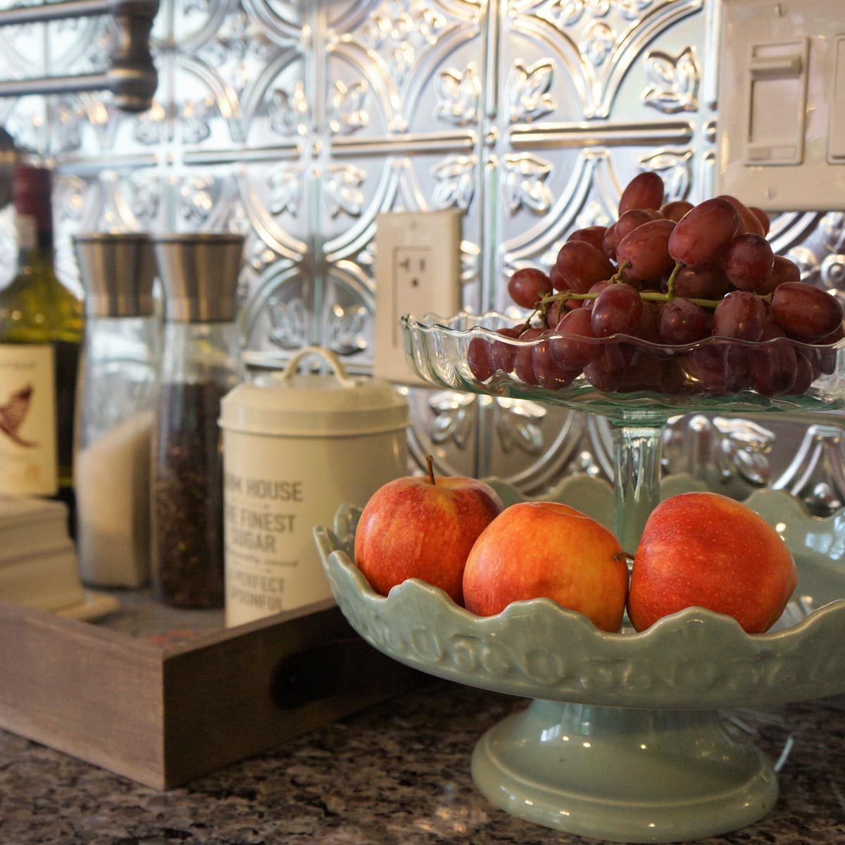 A glass fruit stand displays grapes above a green dish of apples, set against a patterned metallic backsplash in a kitchen environment.