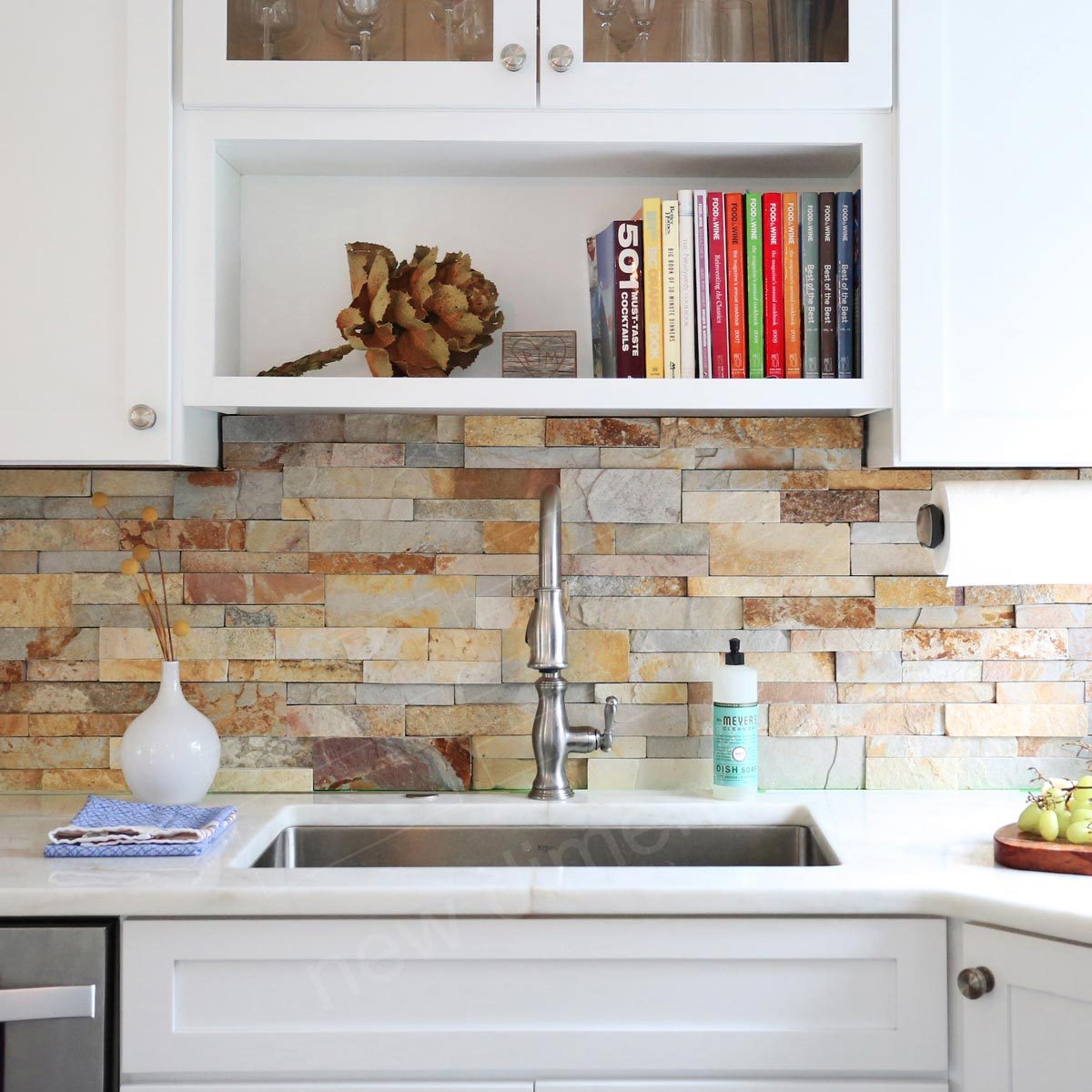 A stainless steel faucet streams water into a white sink on a countertop, with a textured stone backsplash, books on a shelf, and decorative items above.