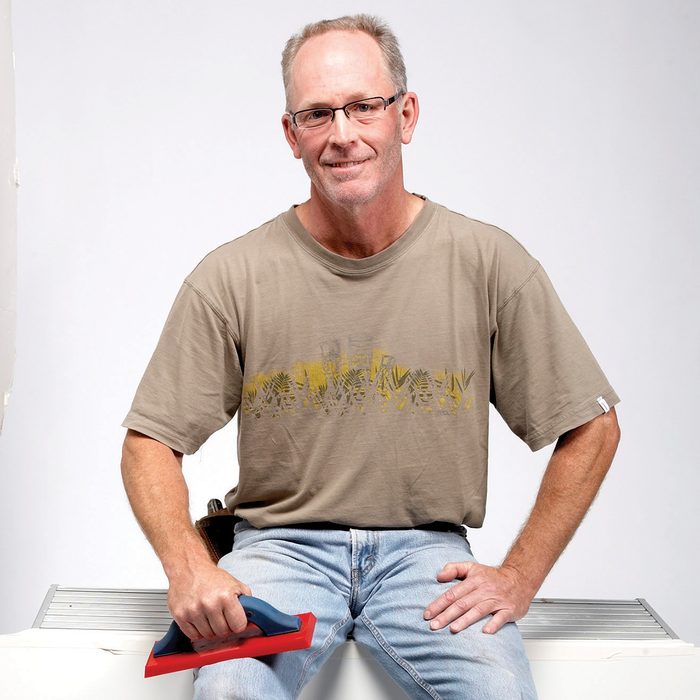 A man sits on a surface, holding a tool with a blue handle and red base, smiling against a simple, light background. Tile Expert Dean Sorem.