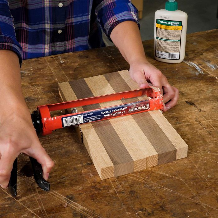 A person uses a caulking gun to apply adhesive on a wooden cutting board on a workbench, with glue nearby for assembly.
